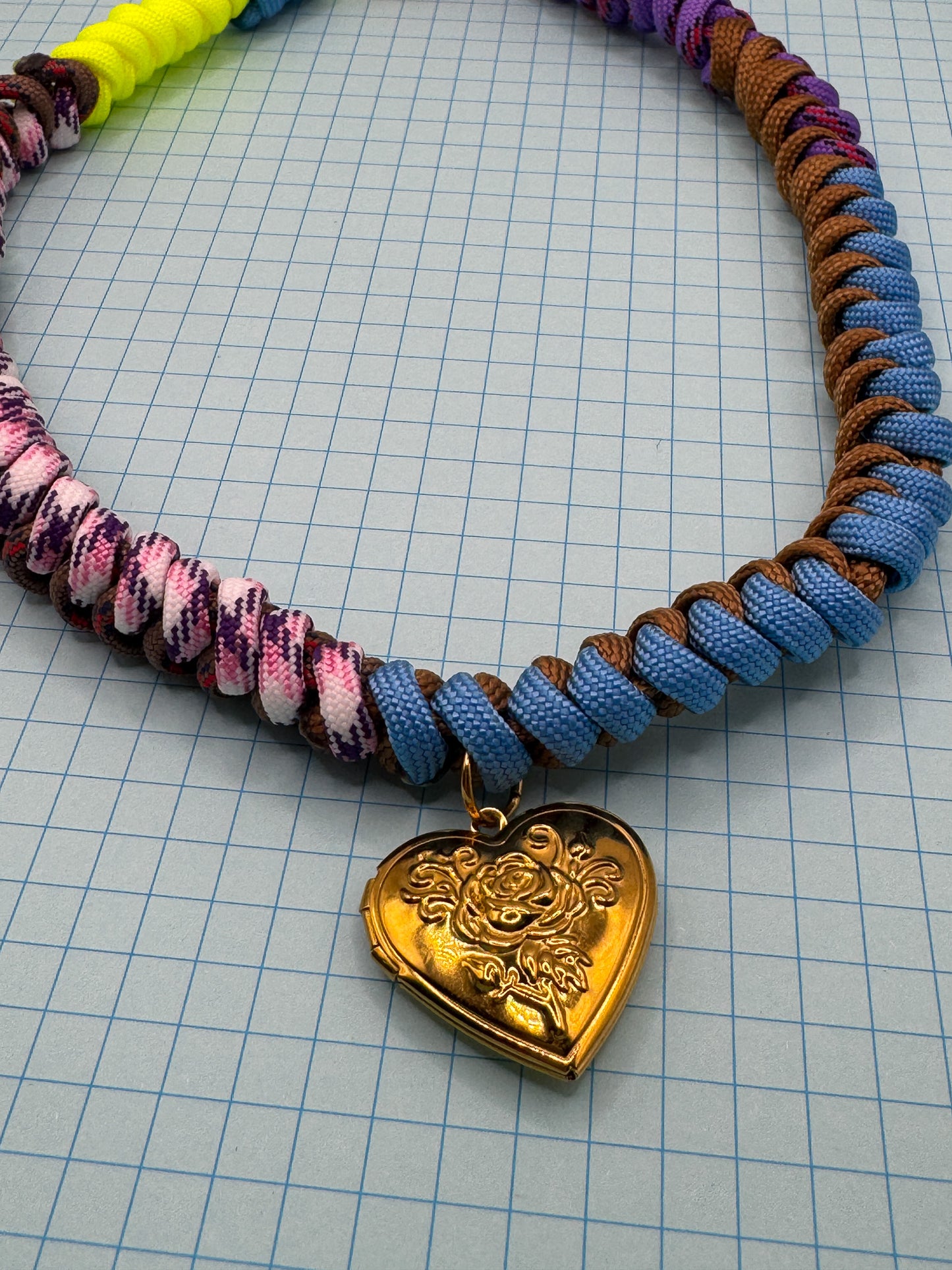 Colorful paracord necklace with a gold heart-shaped pendant on a grid background